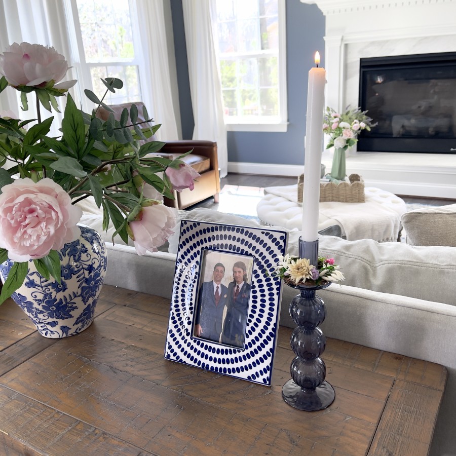 DIY candle ring with grapevine wreath, dried flowers and moss on a glass blue candle holder with white taper candle. Candle ring and candle holder are placed on a brown wood sofa table with a blue and white container of pink peonies and a blue and white ceramic frame with two men in a photograph .