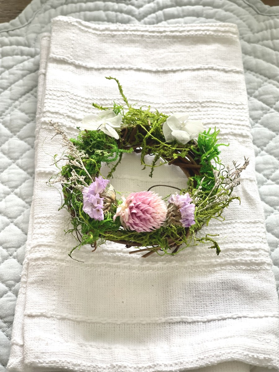 DIY natural mini wreath using 3" grapevine wreath, dried flowers and moss placed on a white cloth napkin with a light blue quilted placemat.