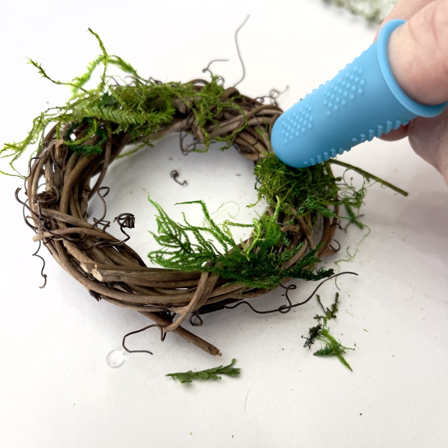 Moss added to a 3-inch grapevine wreath. The person is using silicone finger thimble to protect from hot glue.