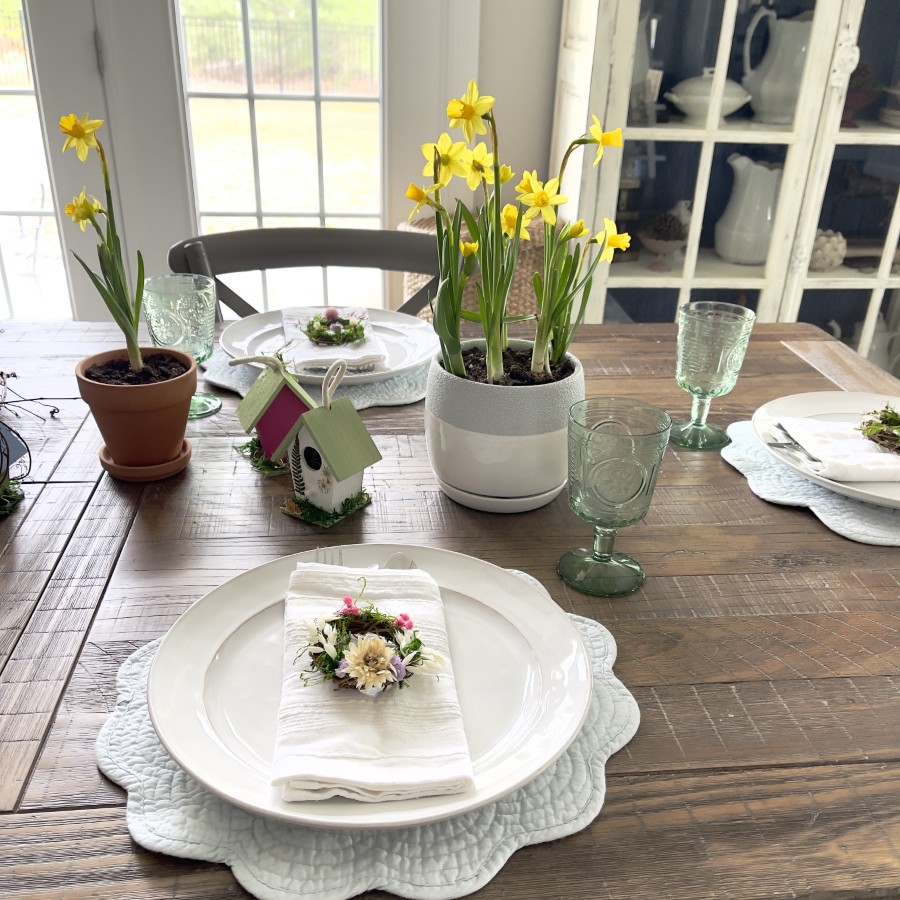 Light blue quilted placemat with a white plate, white cloth napkin with silverware and with a 3" wreath with moss and dried flowers. Green glassware, yellow daffodils in a blue and white ceramic container with decorated birdhouses.