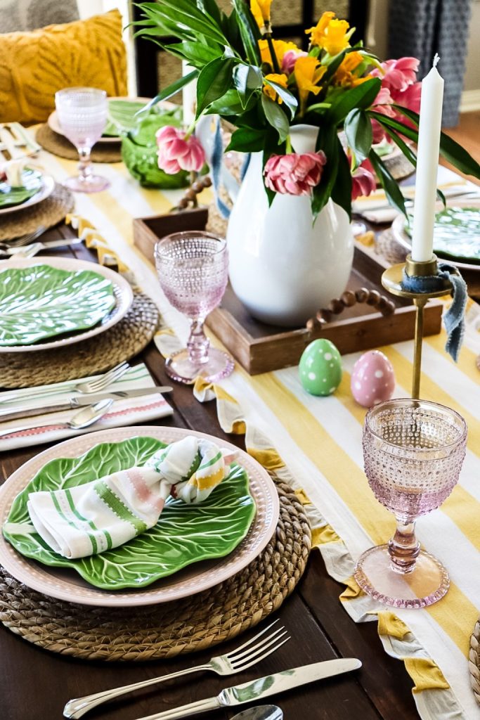 Spring Easter table setting with pink, yellow and green accent colors, cottage leaf plates with striped pastel napkins. Floral arrangement of a white vase with various flowers of pink and yellow.
