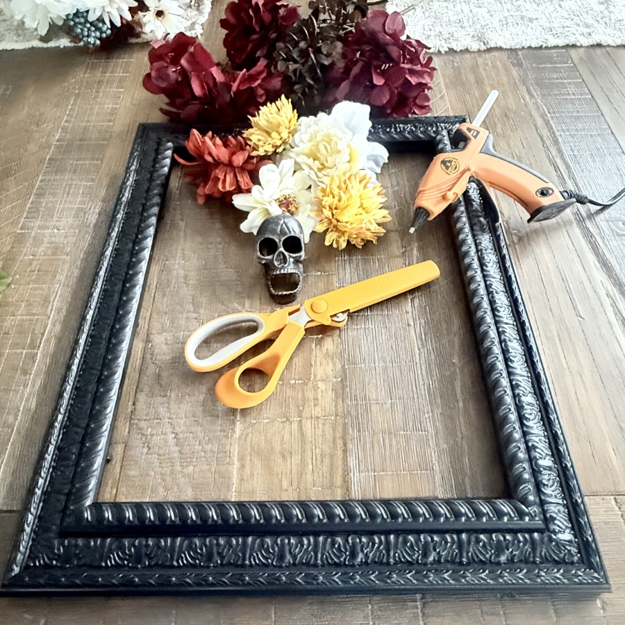 A large black ornate frame with assortment of faux fall flowers, scissors, metal skull and hot glue gun.